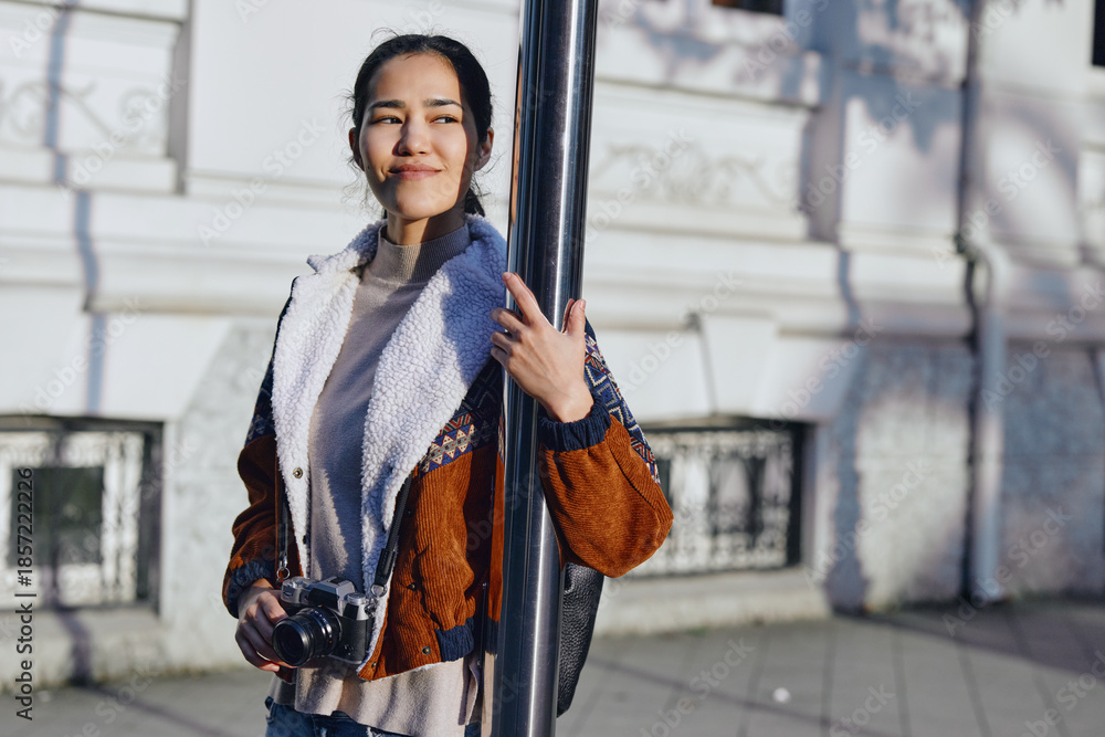 Φωτογραφία Camera in hand, a casual fashion oriented woman leans by a street pole, capturing urban photography and enjoying daylight in a bright outdoor setting with a relaxed smile