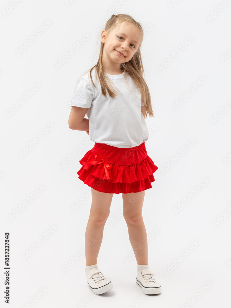 Naklejka premium A happy, smiling 5-year-old child. A girl with long hair is posing and smiling on a white background. She is wearing a T-shirt, a skirt, and sneakers.
