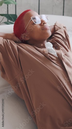 Red haired African American woman with eyeglasses, relaxing on couch wearing casual shirt inside modern living room for leisure and style showing relaxed chill mood and cozy comfortable interior