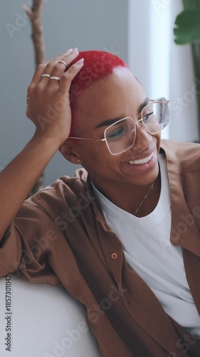 Smiling African American woman with glasses and red hair relaxing on sofa in casual outfit indoor lifestyle with plants and light happy relaxed look and cozy modern home atmosphere
