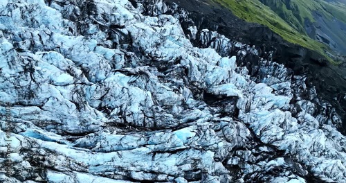 Oblique drone flight along an Iceland glacier edge, showing blue ice blocks with black volcanic ash streaks beneath a green mountainside—dramatic natural climate landscape.