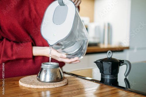 Obraz na płótnie Young woman pours water from filter pitcher into base of moka pot on wooden kitchen island