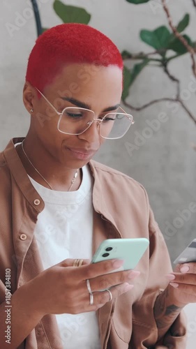 African American female with red hair looks at their phone while holding a card in a contemporary setting. They focus on the devices in their hands and interact with them.