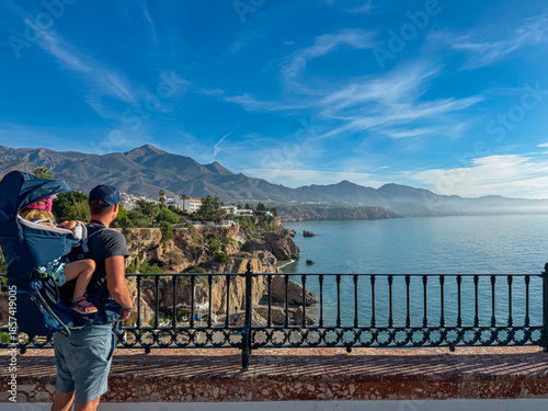 Active father carrying a toddler in a hiking backpack stands by a railing admiring scenic coastline and mountain views in Nerja, Spain, enjoying a family summer vacation on the sunny Costa del Sol.