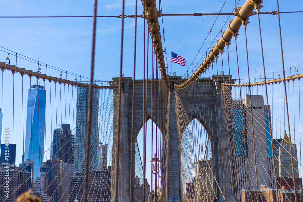 Fototapeta premium View through Brooklyn Bridge cables toward the Manhattan skyline and One World Trade Center with the American flag on the tower.