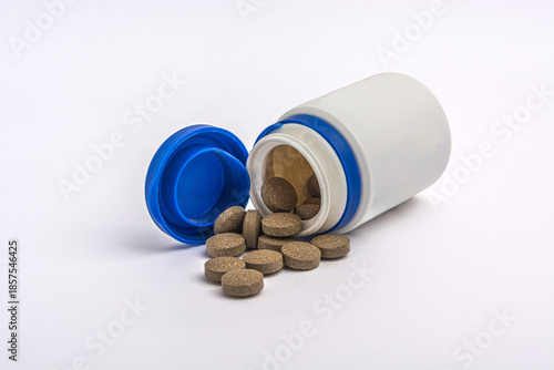 Brown herbal pills spilling out of a white plastic bottle on white background.