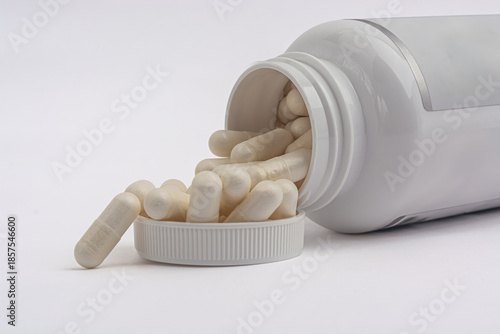 White pill bottle with white capsules spilling into the cap on white background