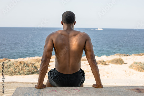 Muscular man performing bench dips by the sea. Shirtless African American athlete doing triceps dips exercise on concrete bench during outdoor workout by the ocean.