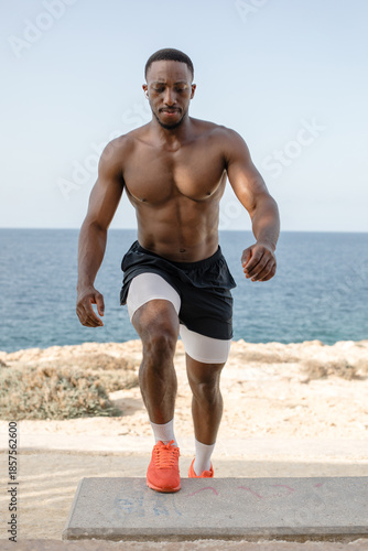 Man doing step-up exercise on bench by the sea. Athletic man performing step-up exercise on a concrete bench along the seaside during outdoor workout.
