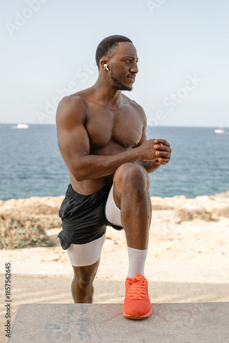 Athletic man performing step-up exercise on concrete bench by the sea. Fit muscular man training legs and balance while doing step-up exercise outdoors on a concrete bench near the ocean.