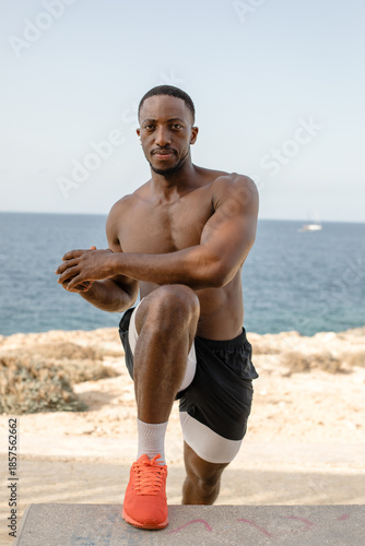 Confident athletic man doing step-up workout outdoors by the ocean. Muscular man exercising outdoors, performing step-up movement on a concrete bench with sea in the background.