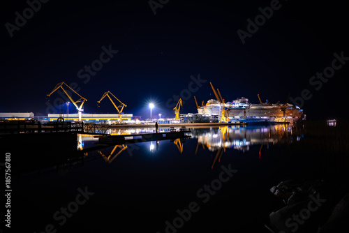 Night view of a massive cruise ship under construction in a modern shipyard with industrial cranes and water reflections.