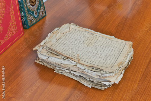 Fotografi Stack of aged pages with Arabic script on wooden table, next to modern Holy Qurans