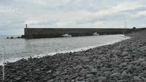 calm waves of the Atlantic ocean rolling on a pebble beach with a breakwater with lighthouse in the background in the harbour of Funchal, Madeira, Portugal