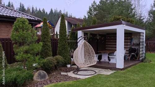 A wooden gazebo with a table, benches and a metal roof and a pine tree are decorated with glowing garlands for Christmas and New Year. There are trees, swings and houses nearby. Cloudy and snowless