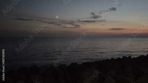 After sunset, the sky with clouds is painted in delicate tones. The moon rose above the horizon. There is a moonlight path and light ripples on the water. There are rocks on the seashore. Evening