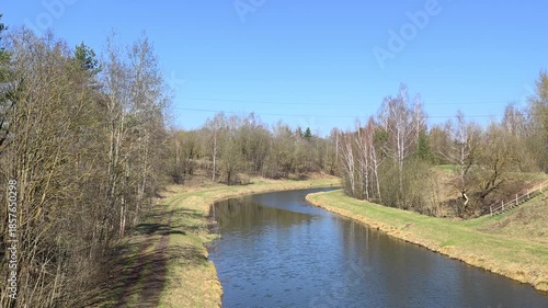 The river flows between grassy banks with a ladder and a walking path. Willow, birch, spruce and pine trees grow nearby. The sky and trees are reflected in the high water. Sunny spring weather