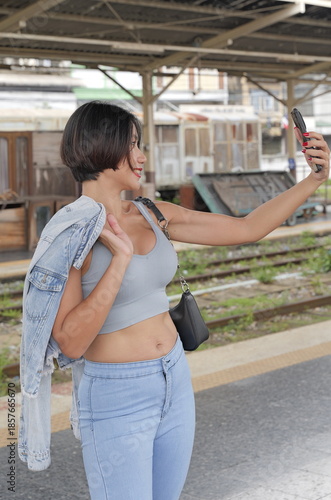 Young woman taking a selfie on a train platform, wearing a light blue outfit with a denim jacket draped over her shoulders, smiling in a candid moment