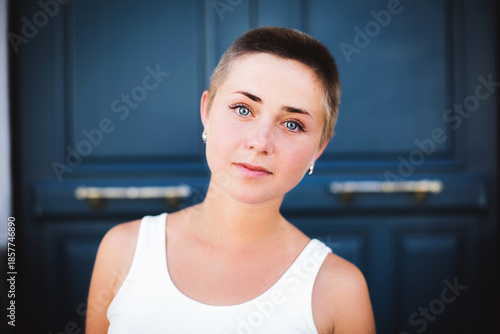 Young woman with short buzz cut hair and piercing blue eyes, tilting her head slightly while looking at the camera against a dark blue door background