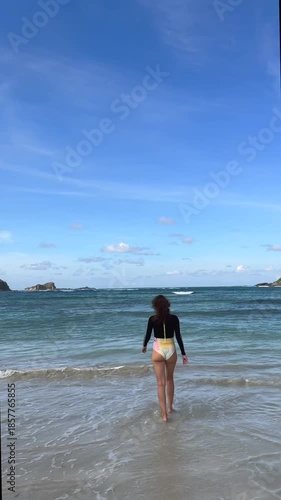 Beautiful young woman wearing a colorful one piece swimsuit walking into the calm turquoise ocean on a sunny day, enjoying her tropical vacation and looking back at the camera