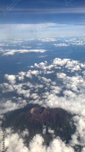 Stunning aerial view from an airplane window of the Agung volcano emerging from the clouds in Bali, Indonesia, with the vast ocean stretching towards the horizon under a clear blue sky