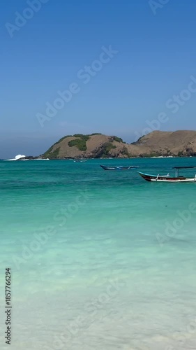 Gentle waves lapping on a pristine white sand beach with traditional Indonesian fishing boats floating on the clear turquoise sea under a bright blue sky, showcasing a serene tropical paradise