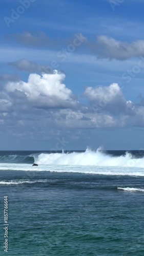 Vibrant turquoise wave crashes and foams over the ocean surface against a backdrop of a clear blue sky dotted with fluffy clouds, capturing a serene yet captivating seascape in slow motion