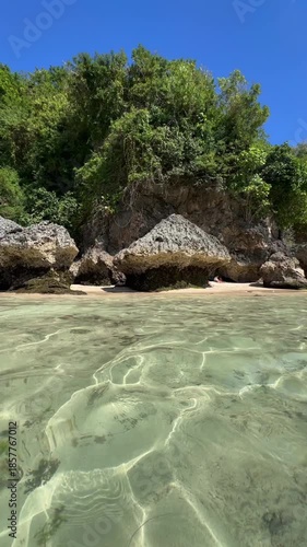 Idyllic summer holiday view of a secluded tropical beach with crystal clear water, sandy seabed, rock formations, and lush green vegetation on a sunny day with a pristine blue sky