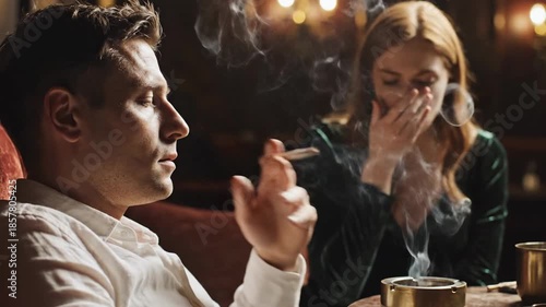 A man holds a cigarette that emits smoke slowly between his fingers while sitting beside a woman who covers her nose with her hand in a dimly lit indoor setting.