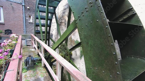 Old water mill exterior with large green metal water wheel, wooden walkway, stone walls, and industrial heritage architecture, no people, overcast daylight, traditional milling technology