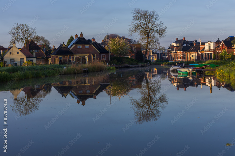 Obraz premium Perfect reflection of the Dutch town Bedum on the canal surface. Traditional brick houses, boats and spring trees in calm morning light