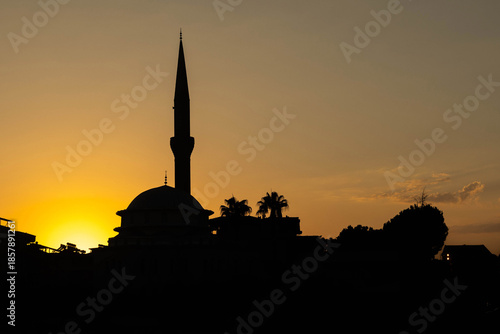 Silhouette of mosque with minaret against vibrant sunset sky. Traditional islamic architecture at golden hour. Time for azan prayer call. August, Mediterranean, Turkey.