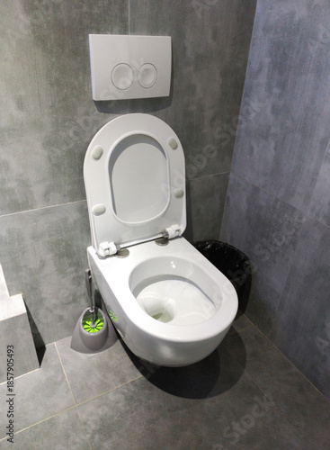 A modern wall-hung toilet with an open lid installed in a gray-tiled bathroom. A dual flush button is mounted on the wall, along with a toilet brush and trash can. A bathroom vanity