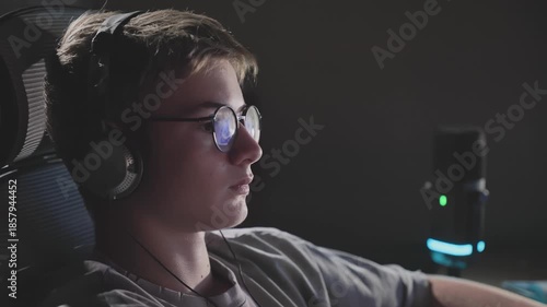 Handheld cinematic shot with subtle camera shake of teenage boy in glasses and headphones at computer, dramatic backlighting, focused on screen activity