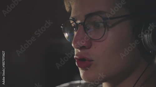Close-up of focused teenage gamer boy wearing glasses and headphones playing online video game and talking to teammates