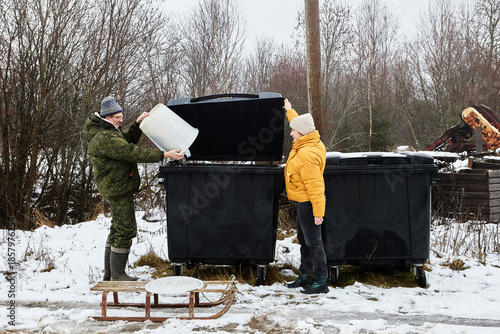 Rural resident empties household trash from waste pail into communal refuse bin at village dump site while spouse keeps cover open.