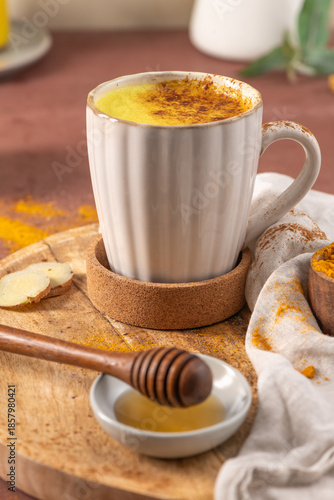 Cozy lifestyle shot of a steaming turmeric latte in a white mug topped with cinnamon with fresh ginger honey and golden spices. Authentic Golden Milk Turmeric Latte Spiced Drink