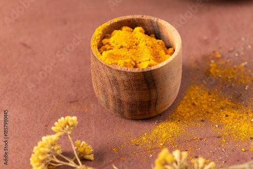 Vibrant yellow turmeric powder fills a small rustic wooden bowl on a warm terracotta surface surrounded by delicate dried flowers and scattered spices