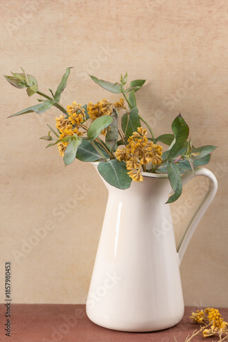 Vintage white enamel pitcher with dried yellow flowers and eucalyptus leaves set against a warm textured background creating a rustic aesthetic