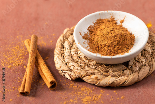 An earthy food photography composition featuring aromatic ground cinnamon and whole quills on a rustic braided coaster against a textured reddish brown background ideal for culinary and baking themes