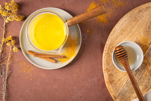 High angle view of a warm turmeric latte in a ceramic mug and glass pot with fresh ginger root honey cinnamon and scattered spices on a rustic tabletop