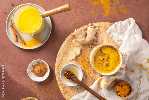 High angle view of a warm turmeric latte served in a ceramic cup and glass pot alongside fresh ginger root honey cinnamon and scattered spices on a rustic tabletop