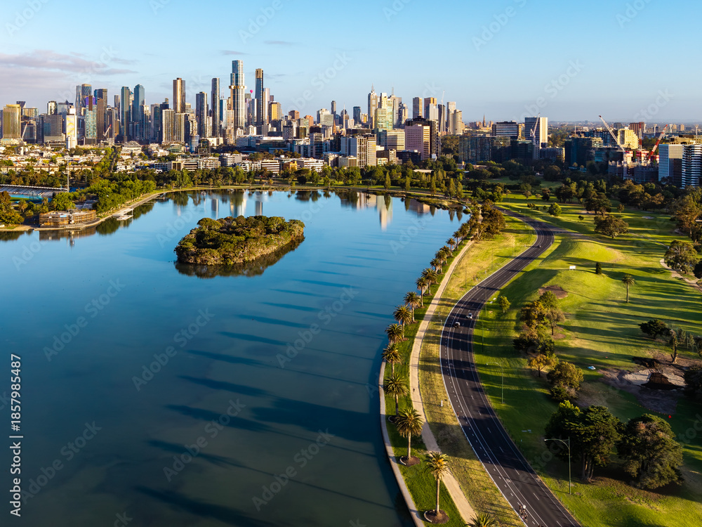 Obraz premium Morning aerial panorama of Melbourne's modern skyline reflecting in Albert Park Lake with vibrant green parklands and pathways.