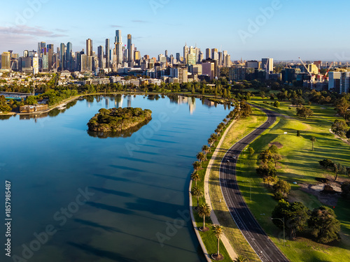 Morning aerial panorama of Melbourne's modern skyline reflecting in Albert Park Lake with vibrant green parklands and pathways.