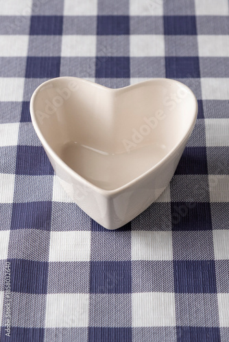 Empty white heart-shaped ceramic bowl on a blue and white checkered tablecloth. Romantic tableware for snacks, desserts or Valentines Day