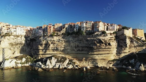 Dramatic view of colorful houses perched atop limestone cliffs overlooking the sea in Bonifacio, Corsica, under a clear blue sky.