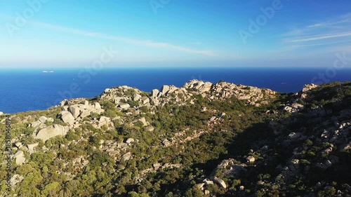 Aerial view of rocky hills covered with green vegetation overlooking the deep blue Mediterranean Sea near Plage de Santa Giulia, Corsica. Clear sky and natural landscape create a serene coastal scene.