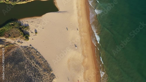 Drone shot of a sandy beach with gentle waves and a lagoon, surrounded by natural vegetation in Algarve, Portugal.