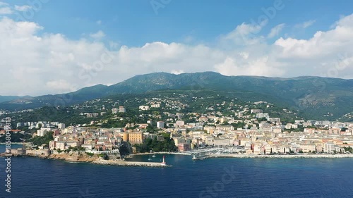 Scenic aerial view of Bastia with colorful buildings along the coastline and lush green mountains in the background, under a bright blue sky.
