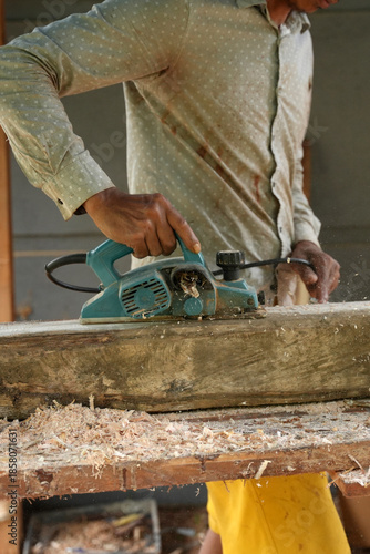 A carpenter shaping a wooden plank using an electric planer, working outdoors as wood shavings scatter, showing a hands on woodworking process in a natural workshop setting.